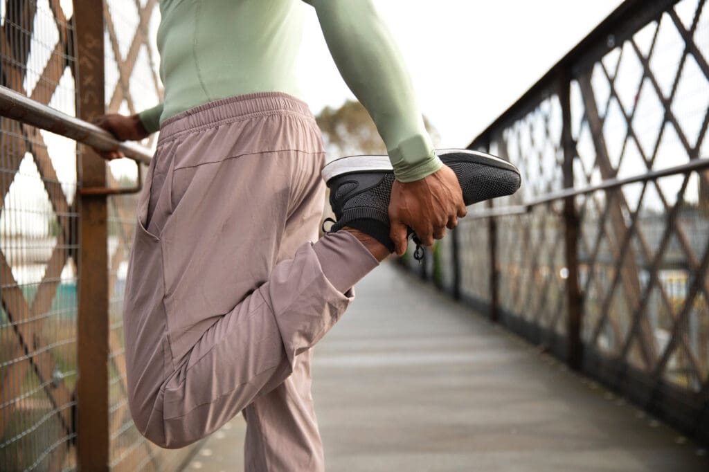 Synthesis Peptides - A person in athletic clothing stretches their leg by pulling their foot toward their back while standing on a bridge, holding onto a railing for balance—highlighting the benefits that research peptides may offer to active individuals.
