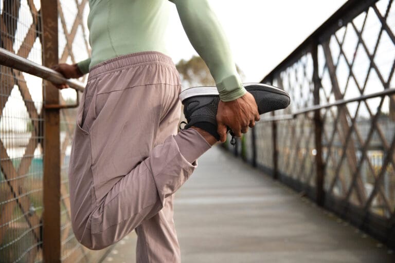 Synthesis Peptides - A person in athletic clothing stretches their leg by pulling their foot toward their back while standing on a bridge, holding onto a railing for balance—highlighting the benefits that research peptides may offer to active individuals.
