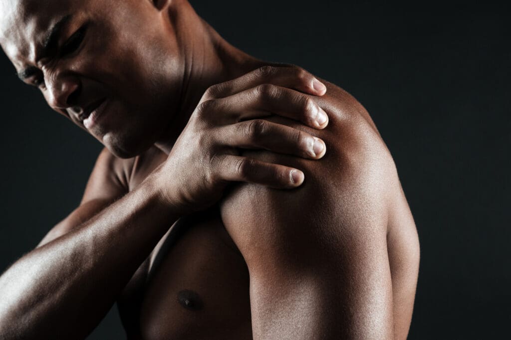 Synthesis Peptides - A man grimacing in pain holds his left shoulder with his right hand, suggesting discomfort or injury, against a dark background—an image often used in discussions about the potential benefits of research peptides.
