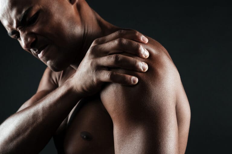 Synthesis Peptides - A man grimacing in pain holds his left shoulder with his right hand, suggesting discomfort or injury, against a dark background—an image often used in discussions about the potential benefits of research peptides.