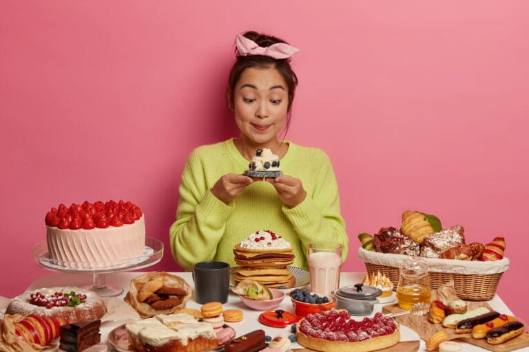 Synthesis Peptides - A woman with a pink bow and green sweater excitedly looks at a cupcake, sitting at a table full of cakes, pancakes, pastries, fruits, and desserts—perfect fuel for anyone curious about the sweetness of research peptides.