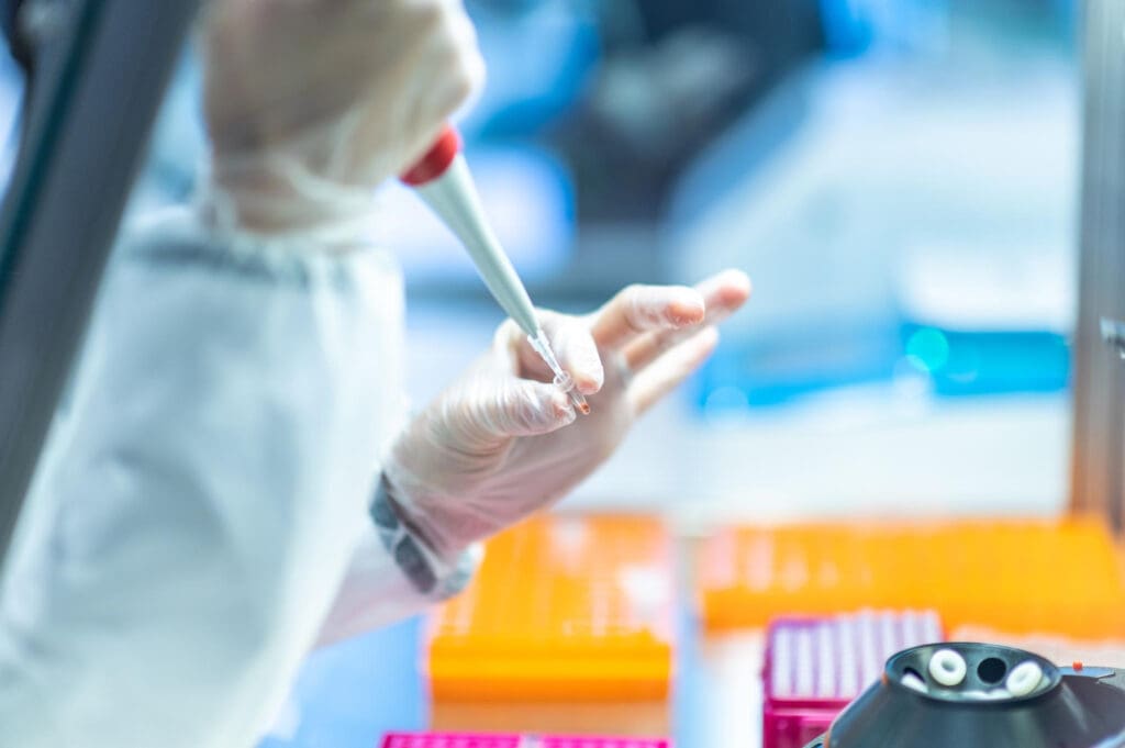 Synthesis Peptides - A scientist wearing gloves uses a pipette to transfer research peptides in a laboratory, with orange sample trays and lab equipment visible in the background.