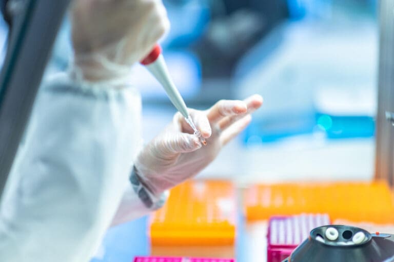 Synthesis Peptides - A scientist wearing gloves uses a pipette to transfer research peptides in a laboratory, with orange sample trays and lab equipment visible in the background.