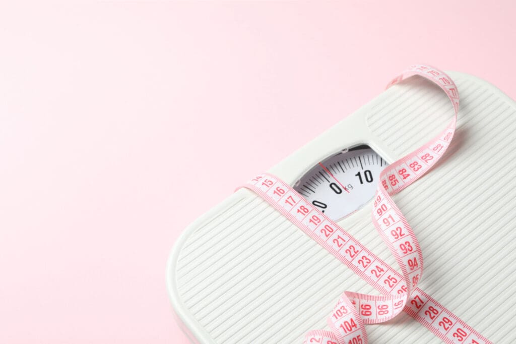 Synthesis Peptides - A white bathroom scale with a pink measuring tape draped over it, set against a light pink background. The scale’s dial is visible, showing numbers, and the tape is loosely wrapped on top—an inviting scene for those interested in peptides and healthy living.