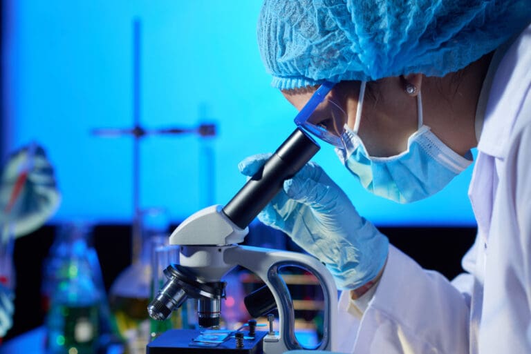 Synthesis Peptides - A scientist wearing a lab coat, gloves, face mask, and hair cover examines research peptides through a microscope in a laboratory filled with glassware and equipment, all illuminated by blue light.