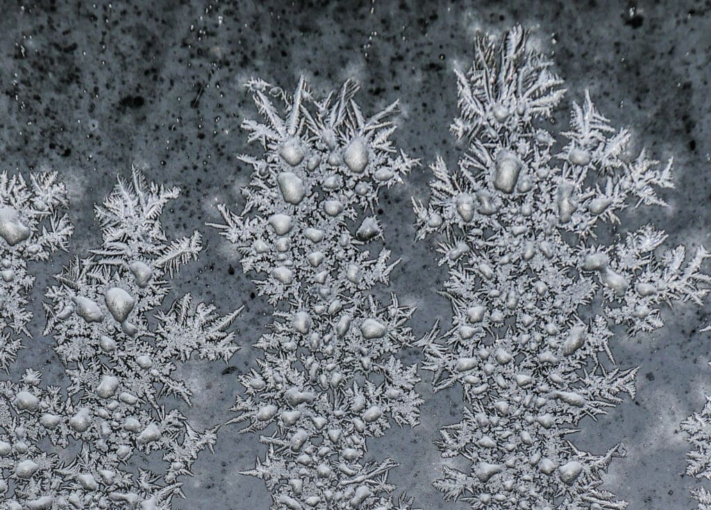 Synthesis Peptides - Close-up of intricate frost patterns and ice crystals on a window, forming dense clusters and delicate, spiky shapes—resembling the complex structures seen in research peptides—set against a dark, mottled background.